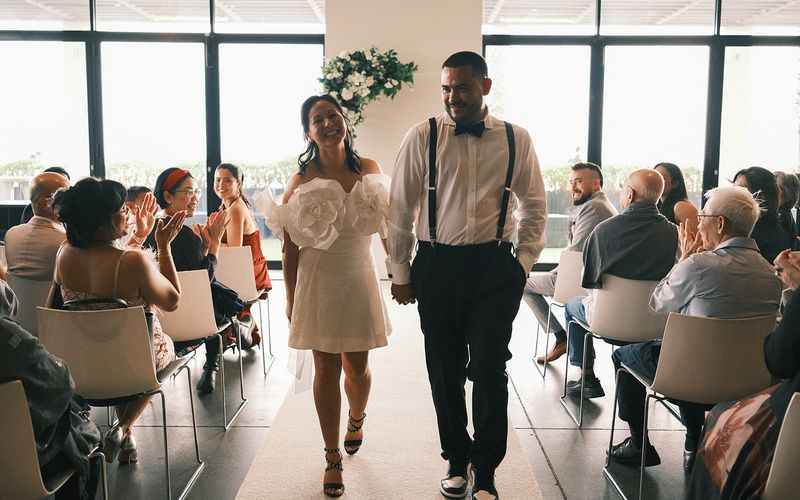 Deb and Mickey walking hand in hand back up the aisle at Encore St Kilda Beach as their guests applaud