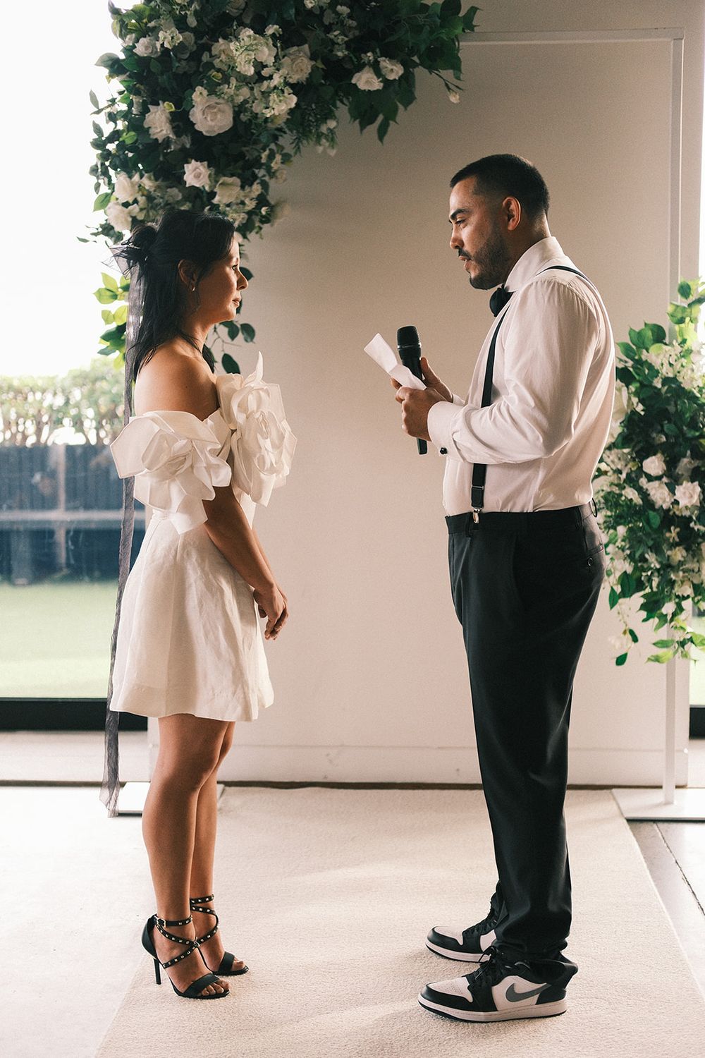 Mickey reading his wedding vows to Deb beside an arrangement of white flowers and greenery