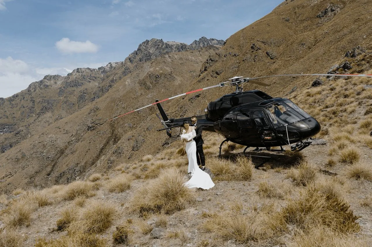 A photo of a couple with the helicopter they flew to the mountain top in Queenstown New Zealand with photographed by George Bowden for the Elopement Collective with wedding celebrant Josh Withers from Tasmania ⌘