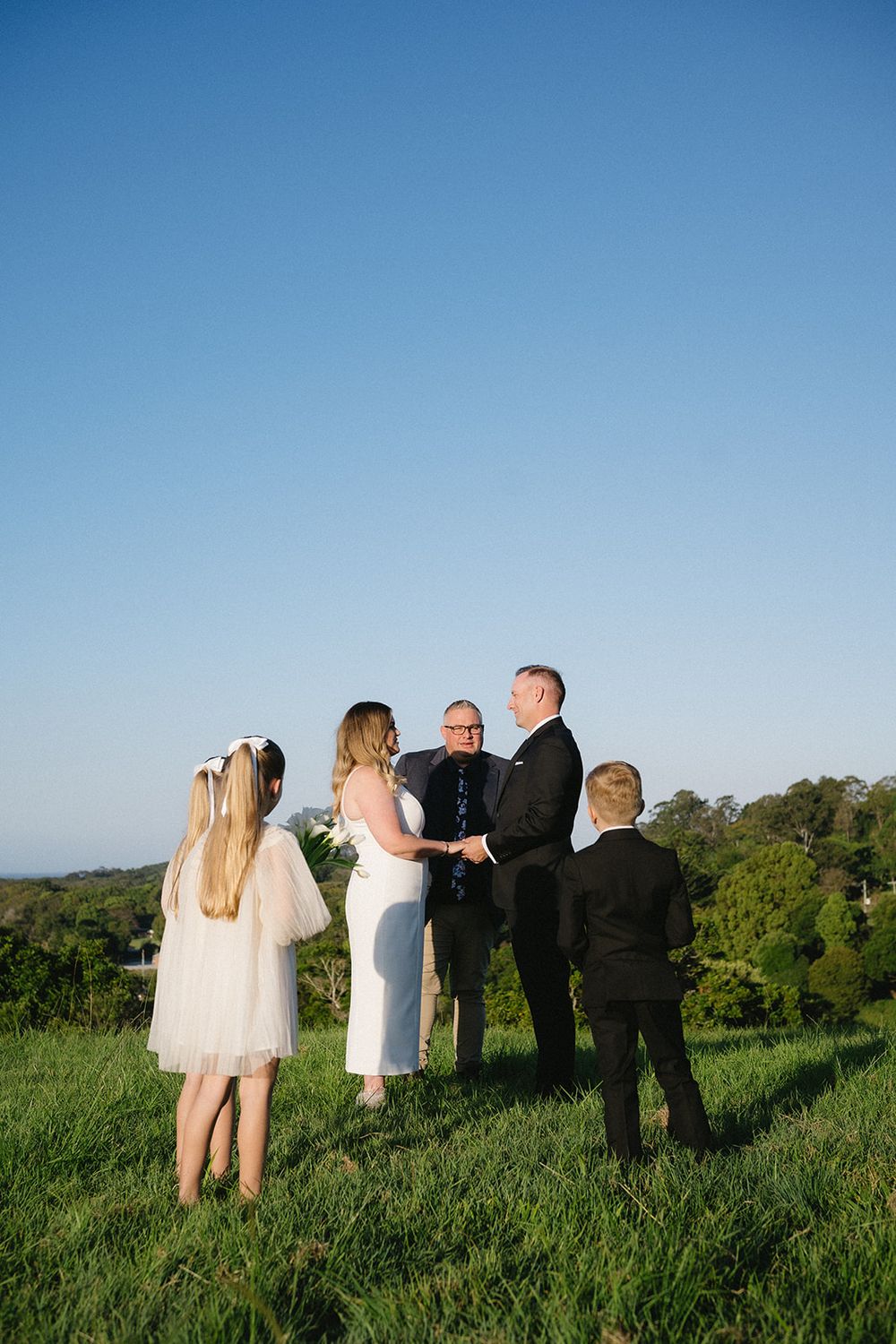 Libby and Daniel holding hands with Josh Withers beside them on a grassy hill while two young guests watch