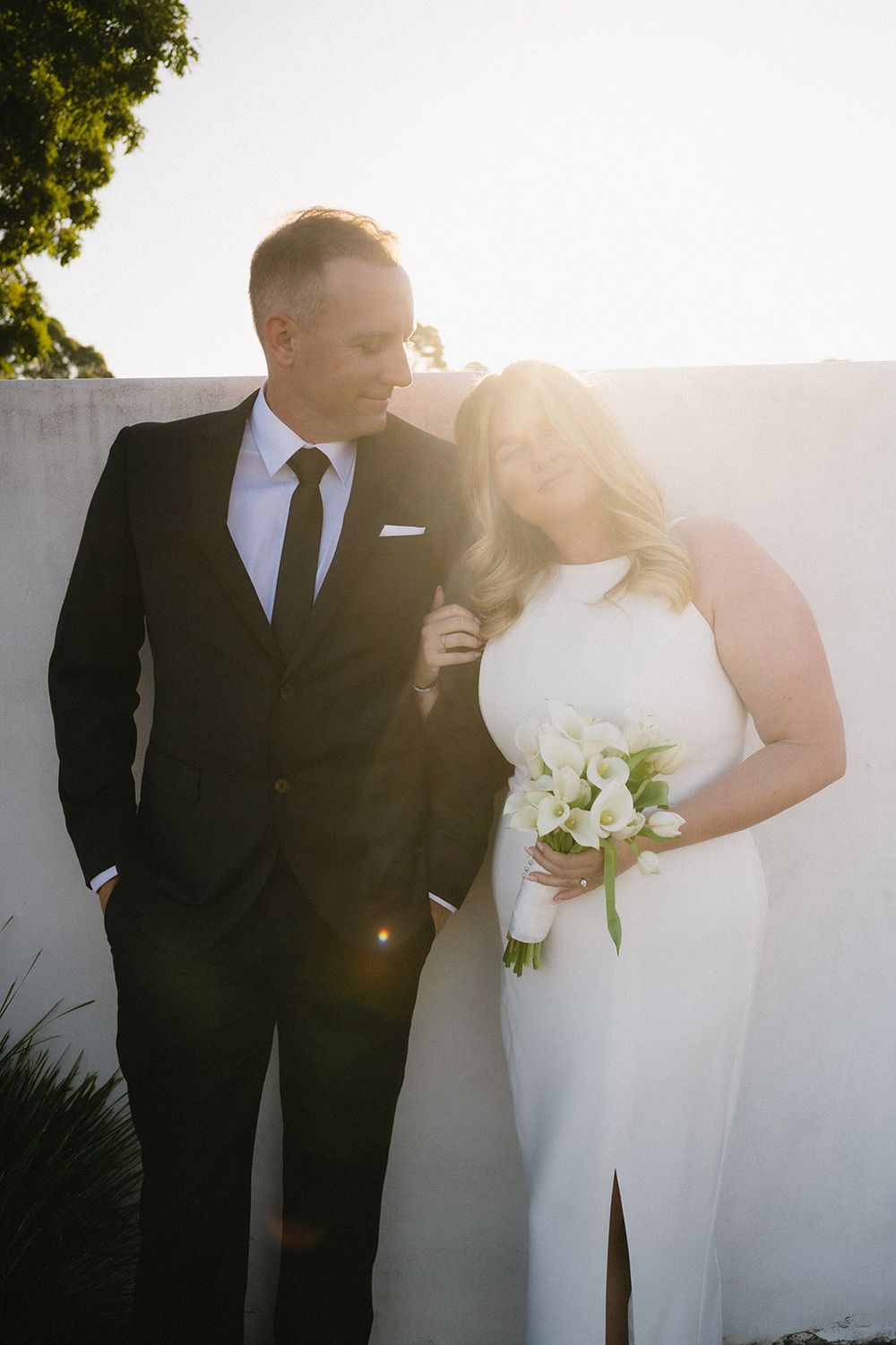 Libby holding her white bouquet beside Daniel against a pale wall in the afternoon sunlight