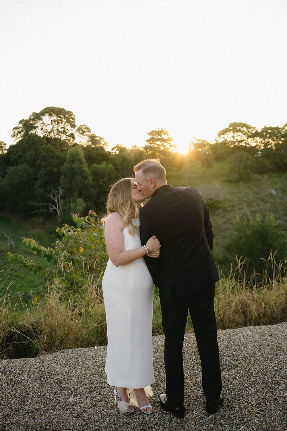 Libby and Daniel kissing on a gravel path with the setting sun shining through the trees behind them