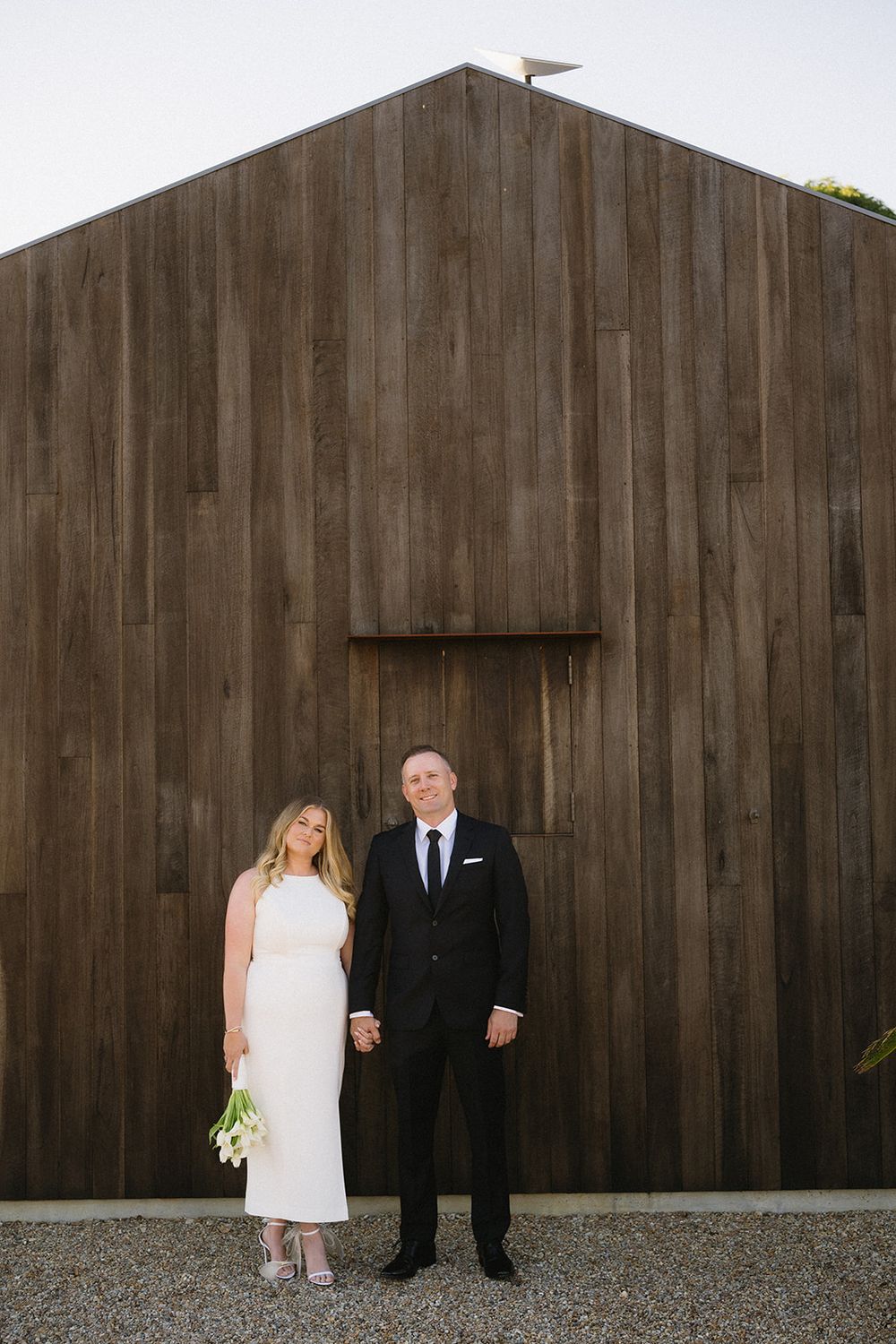 Libby and Daniel holding hands in front of a tall timber wall, with Libby carrying a white calla lily bouquet