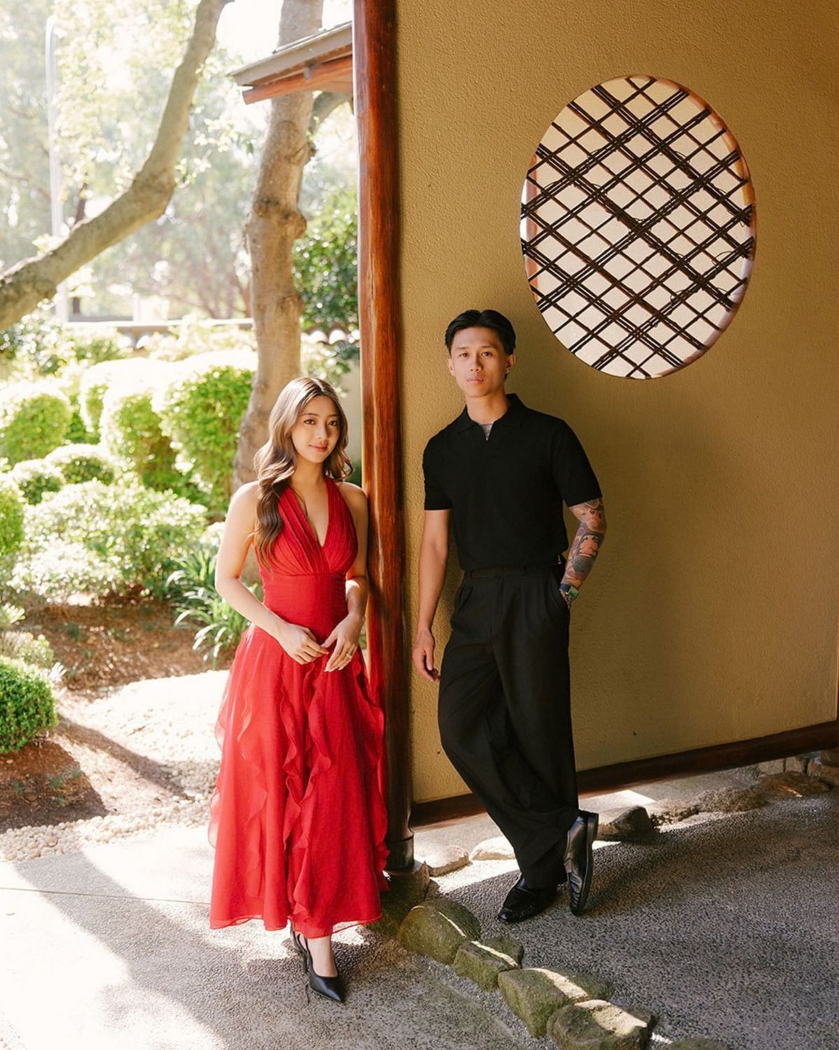 Sherrie in a red dress and Jeremy in black standing beside a circular lattice window in a garden